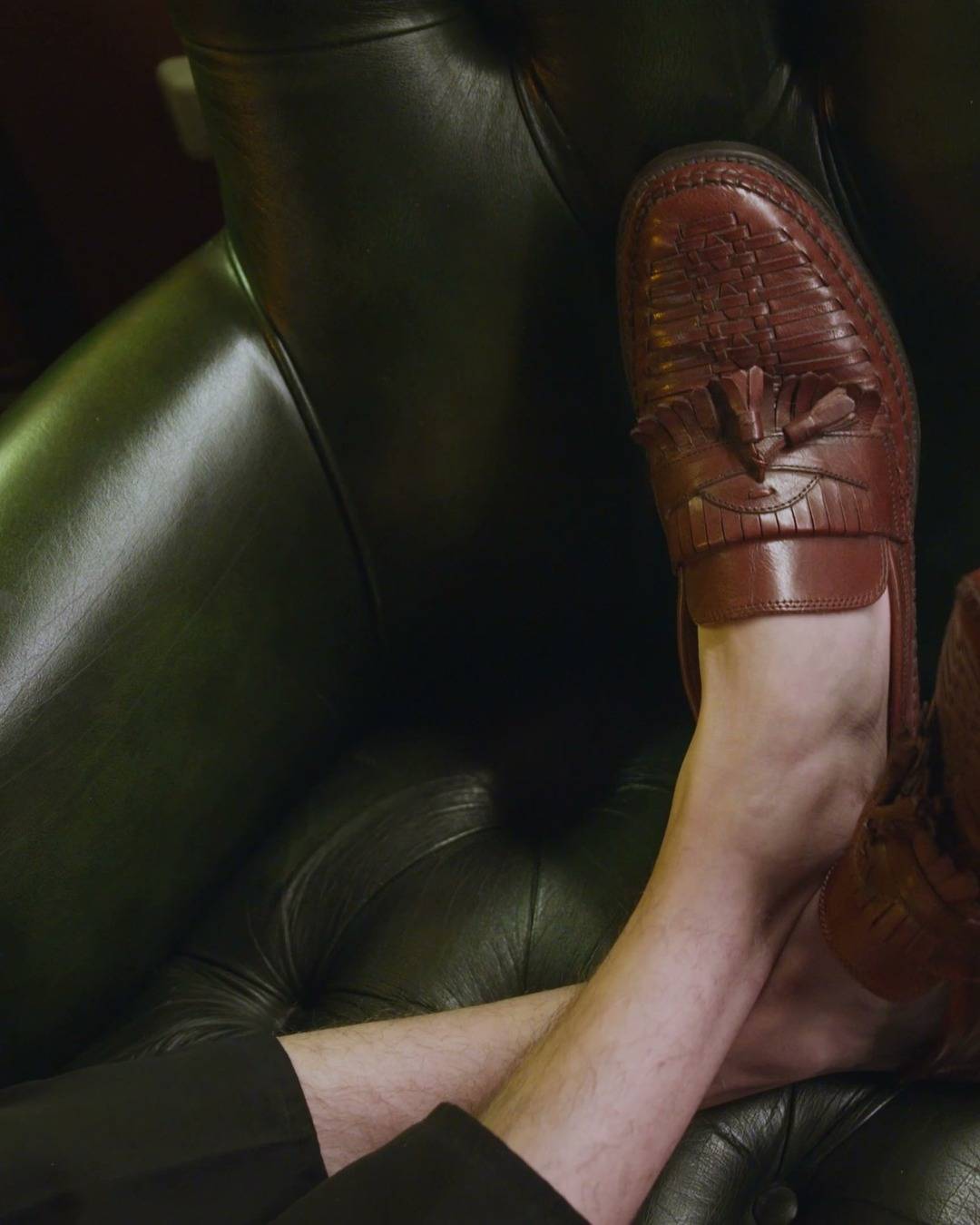 Mocasín de cuero marrón con borlas sobre un sillón Chesterfield verde.